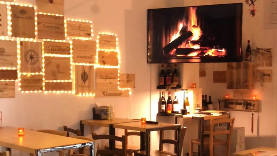 La sala: una parete di cassette di legno da vino contornate da lucine calde, tavoli di legno con le candele accese e una mensola di bottiglie.
