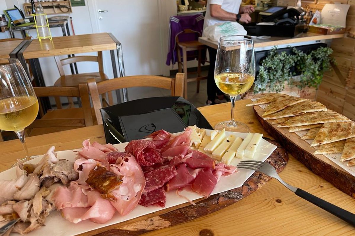 Un tagliere di legno con salumi affettati e formaggi, accanto a spicchi di piadina calda e due calici di vino bianco, su un tavolo di legno del locale.